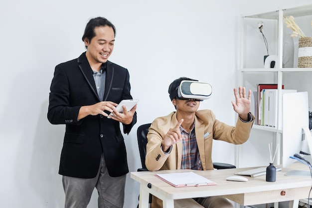 Asiatische junge Arbeiter, die im Büro eine Simulation mit VR-Brille und Tablet-Smartphone haben.