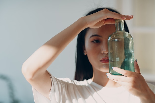 Asiatische junge erwachsene frau hält grüne glasflasche vorderseite mit tagsonnenlicht drinnen