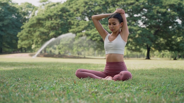 Asiatische junge Frau, die auf dem Gras sitzt und Yoga in der Kuhhaltung im Stadtpark mit dem Hintergrund der großen Bäume praktiziert Rückansicht von Frauen, die an einem sonnigen Tag Yoga im Freien praktizieren