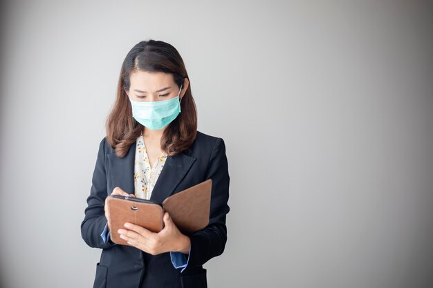 Asiatische junge Frau, die eine Tablette verwendet, um von zu Hause aus zu arbeiten und eine Antivirenmaske zu tragen, um andere vor Coronavirus zu schützen