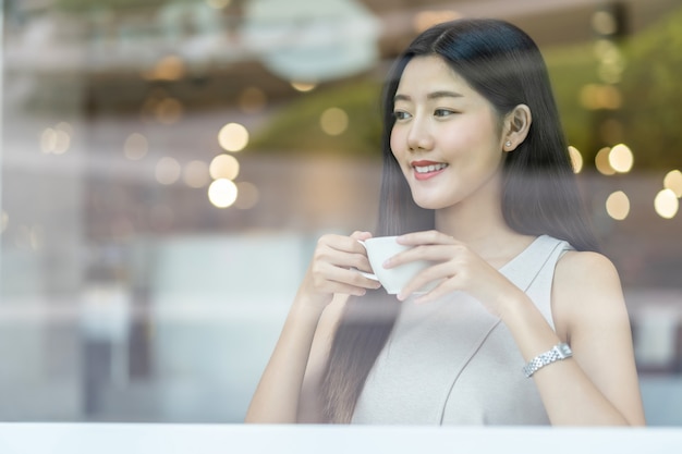 Asiatische junge Frau, die einen Tasse Kaffee in der modernen Kaffeestube oder im Coworkingraum neben Fensterspiegel, Japaner, Chinesen, koreanischem Lebensstil und Alltagsleben, Unternehmerkonzept hält