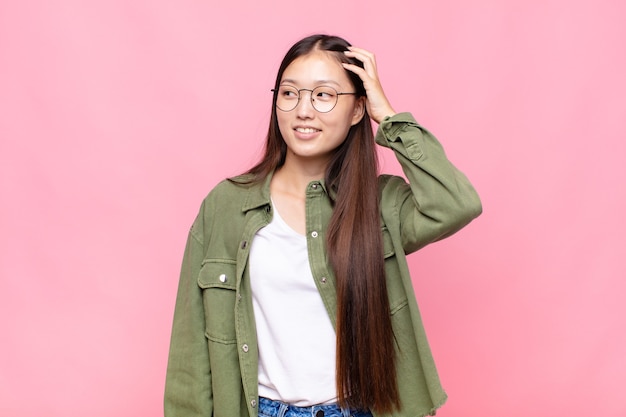 Asiatische junge Frau, die fröhlich und beiläufig lächelt und Hand an Kopf mit einem positiven, glücklichen und selbstbewussten Blick nimmt