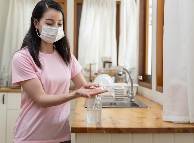 Asiatische junge Frau, die Gesichtsmaske oder Schutzmaske trägt, die zu Hause arbeitet und ihre Hände mit Desinfektionsgel während des Ausbruchs von Coronavirus oder Covid 19 reinigt.