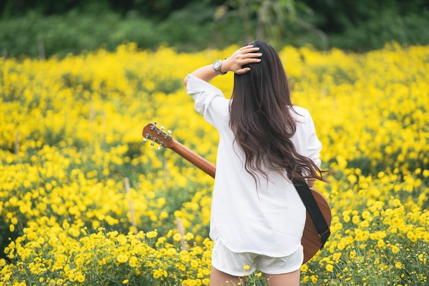 Asiatische junge Frau, die Gitarre spielt und Musik im Park singt, asiatische Frau, die Gitarre im gelben Blumengarten spielt