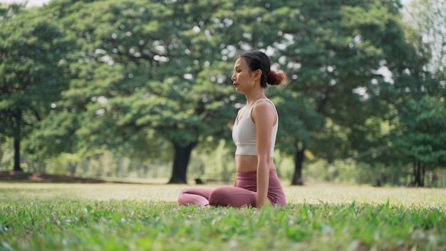 Foto asiatische junge frau, die im lotussitz auf dem gras sitzt und die hände draußen im stadtpark mit dem hintergrund der großen bäume hebt rückansicht von frauen, die an einem sonnigen tag yoga im freien praktizieren