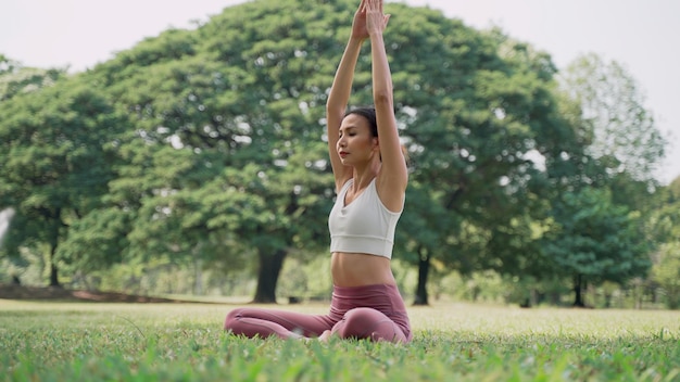 Asiatische junge Frau, die im Lotussitz auf dem Gras sitzt und die Hände draußen im Stadtpark mit dem Hintergrund der großen Bäume hebt Rückansicht von Frauen, die an einem sonnigen Tag Yoga im Freien praktizieren