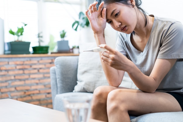 Foto asiatische junge frau, die mit coronavirus oder covid-19 krank ist und hohe temperatur hat, sitzt zu hause auf dem sofa.