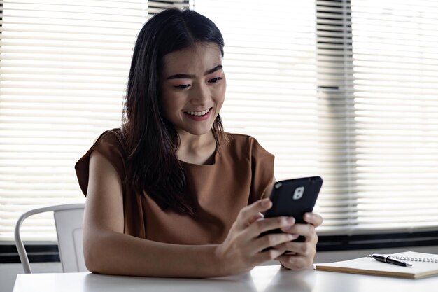 Asiatische junge Frau, die online auf dem Handy mit glücklichem Lächeln im Arbeitszimmer zu Hause sozial spielt. Glückliche Frau, die Online-Internet auf dem Smartphone spielt.