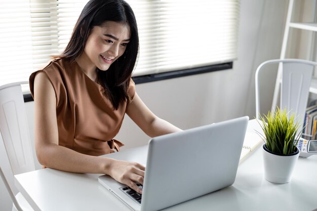 Asiatische junge Frau, die online auf Laptop mit glücklichem Lächeln im Arbeitszimmer zu Hause sozial spielt. Glückliche Frau, die Online-Internet auf Computernotizbuch spielt.