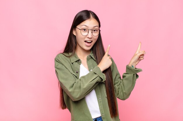 Asiatische junge Frau, die sich freudig und überrascht fühlt, mit einem schockierten Ausdruck lächelt und zur Seite zeigt