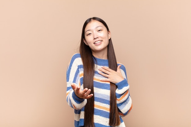 Asiatische junge Frau, die sich glücklich und verliebt fühlt und mit einer Hand neben dem Herzen und der anderen vorne gestreckt lächelt