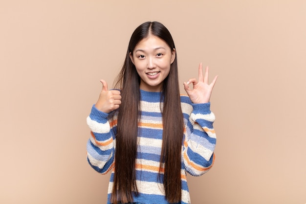 Asiatische junge Frau fühlt sich glücklich, erstaunt, zufrieden und überrascht, zeigt okay und Daumen hoch Gesten, lächelnd