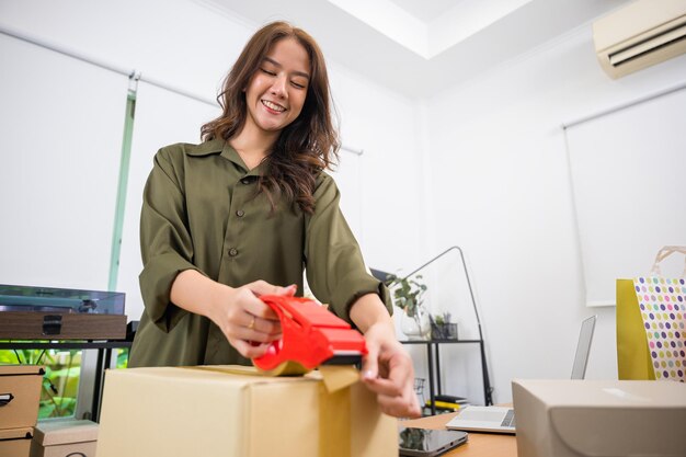 Asiatische junge Frau handverpackt Versandkarton mit Transparentband im Home Office, glückliche Freiberuflerin und Verpackung von Kartonlieferprodukten an Kunden, Startup-Kleinunternehmen, Umzugstag