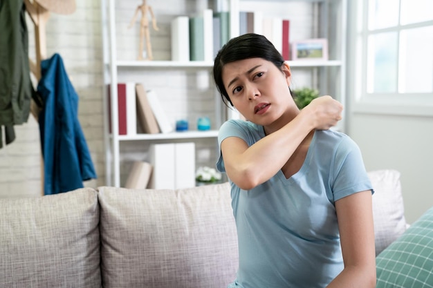 asiatische junge frau mit schulterschmerzen, die auf der couch sitzt und ihre körpermassage fallen lässt, um schmerzhaftes gefühl zu lösen. chinesische schöne Hausfrau, die sich zu Hause auf dem Sofa entspannt. Krankheit Dame überbeansprucht Handy