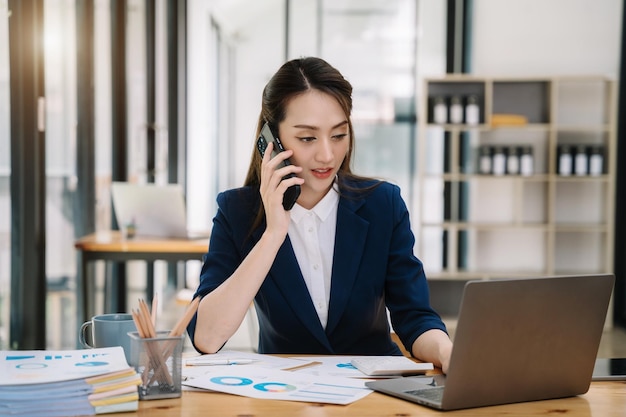 Asiatische junge Frau telefoniert mit Mobiltelefonen und macht sich Notizen auf ihren Notizbüchern, während sie an Laptop-Computern arbeitet. Online-Business-Telearbeitskonzept