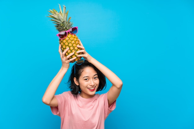 Asiatische junge Frau über dem lokalisierten blauen Hintergrund, der eine Ananas mit Sonnenbrille hält