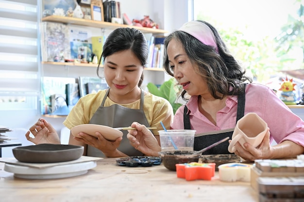 Asiatische junge Frau und gealterte Frau malen gemeinsam eine Keramikplatte in der Werkstatt