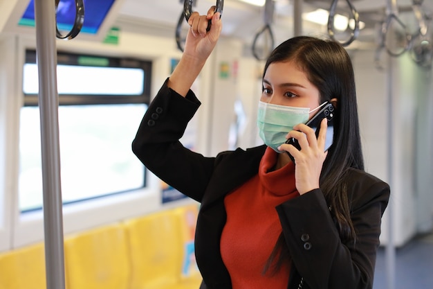 Asiatische junge Geschäftsfrau, die Maskenschutz trägt, um Virus-covid19-Infektion von Leuten im Zug zu verhindern, unter Verwendung der Smartphone-Kommunikation im Zug