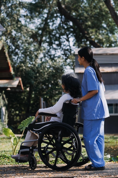 Asiatische junge Krankenschwester im Pflegeheim kümmert sich um einen behinderten älteren Mann. Betreuerarzt bietet Physiotherapie für ältere ältere Patienten an, um das Gehen auf Gehhilfe oder Stock im Hinterhof zu trainieren und zu üben