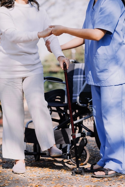 Asiatische junge Krankenschwester im Pflegeheim kümmert sich um einen behinderten älteren Mann. Betreuerarzt bietet Physiotherapie für ältere ältere Patienten an, um das Gehen auf Gehhilfe oder Stock im Hinterhof zu trainieren und zu üben