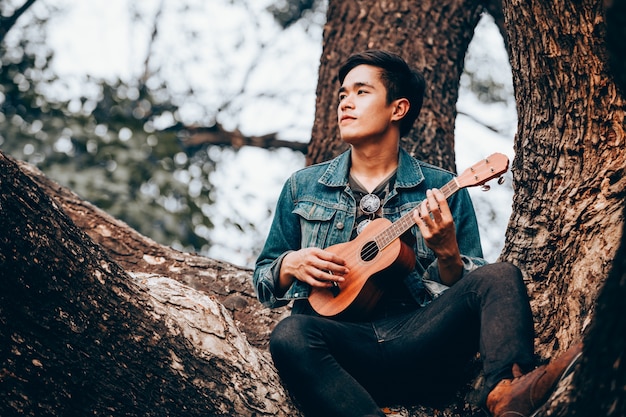 Asiatische junge Männer des Porträts, die auf Baumspielukulele sitzen