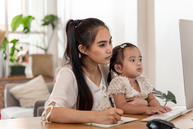 Asiatische junge Mutter, die von zu Hause aus arbeitet und Baby hält, während sie am Telefon spricht und Computer benutzt, während sie Zeit mit ihrem Baby verbringt