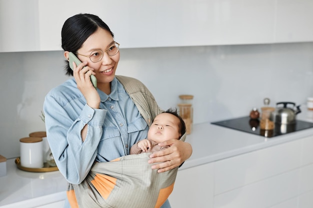 Asiatische junge Mutter hält Baby im Tragetuch, während sie sich auf dem Handy unterhält