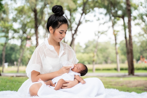 Asiatische junge schöne Mutter, die ihr Neugeborenes hält, schläft und fühlt sich mit Liebe an und berührt sanft, dann sitzt sie auf grünem Gras im Park