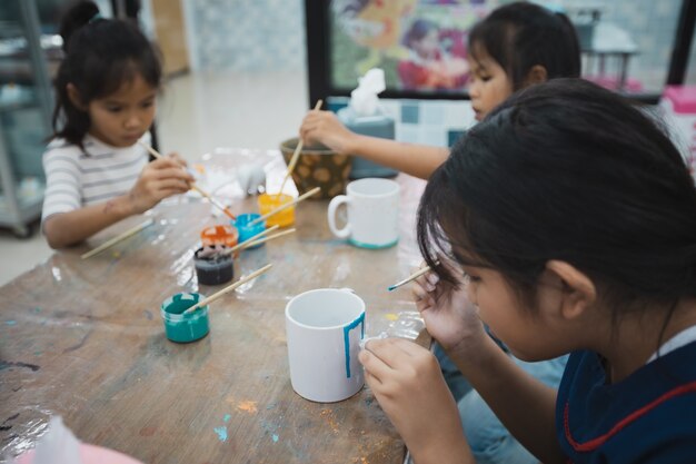 Asiatische Kinder und Freunde konzentrieren sich darauf, mit Spaß zusammen mit Öl auf Keramikglas mit Ölfarbe zu malen. Kreative Aktivitätsklasse für Kinder in der Schule.