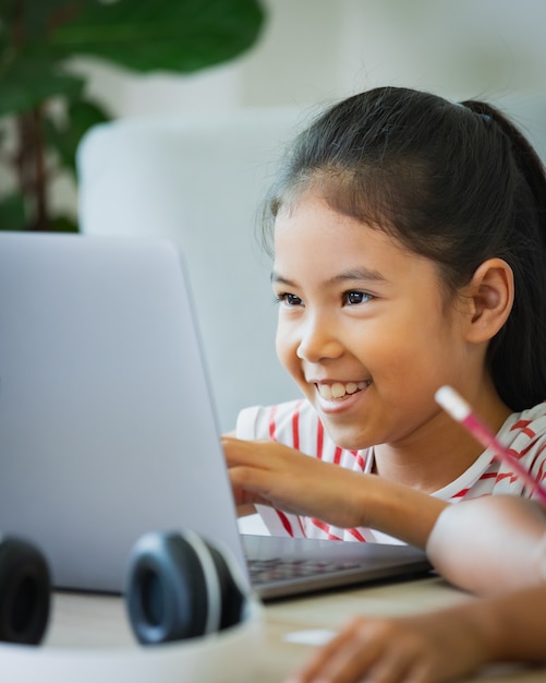 Foto asiatische kindermädchen tragen kopfhörer online mit dem lehrer per videoanruf. das kind unterrichtet während der quarantäne aufgrund der covid-19-pandemie mit computer-laptop.