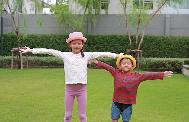 Asiatische kleine Jungen und Mädchen tragen Strohhut und öffnen breite Arme mit Blick auf die Kamera im Garten