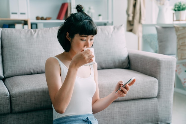 asiatische koreanerin lächelt, chattet per smartphone und genießt morgens die kaffeepause zu hause. Elegante Frau in weißem Tanktop, die eine Tasse Tee trinkt und drinnen soziale Medien auf dem Handy anschaut.