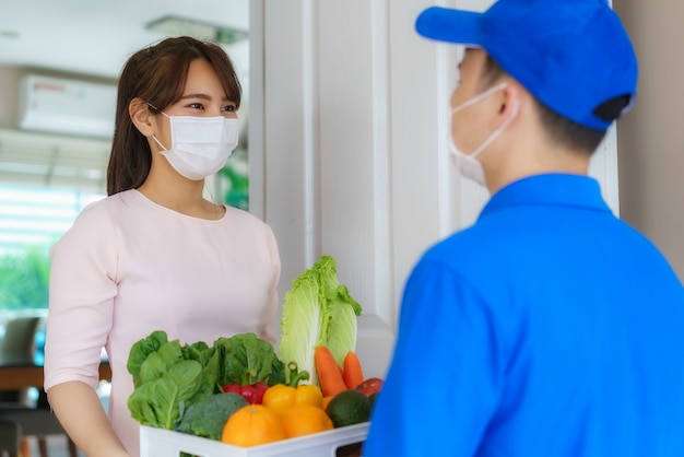 Asiatische Kostümierin, die Gesichtsmaske und Handschuh trägt, erhält Lebensmittelkiste mit Lebensmitteln, Obst, Gemüse und Getränken vom Lieferboten vor dem Haus während der Zeit der Hausisolation.