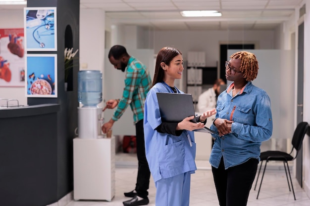 Asiatische Krankenschwester zeigt der Person in der Empfangshalle des Krankenhauses die Krankheitsdiagnose und erklärt die medizinischen Ergebnisse auf dem Laptop. Verschiedene Frauen sprechen im Wartebereich über medizinische Behandlung und Genesung.