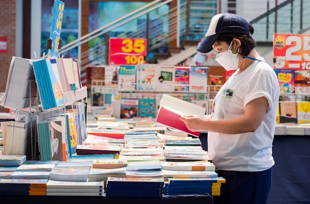 Asiatische LGBT-Frau im lässigen Stil mit Mütze und Schutzmaske, die Bücher im Buchladen auswählt