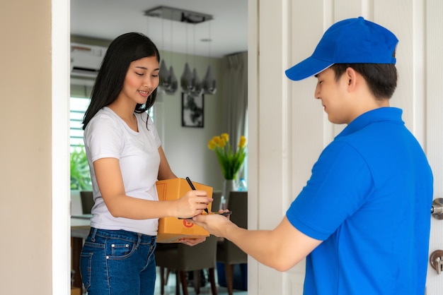 Asiatische Lieferung junger Mann im blauen Uniformlächeln und im Smartphone im Vorderhaus haltend