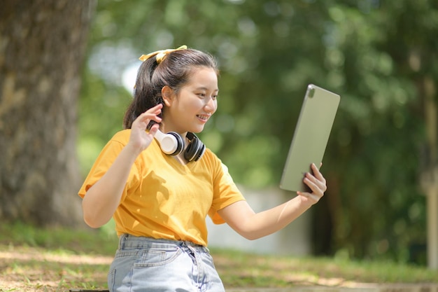 Asiatische Mädchen begrüßen Freunde mit einem Tablet, das ein gelbes Hemd und Kopfhörer am Hals trägt