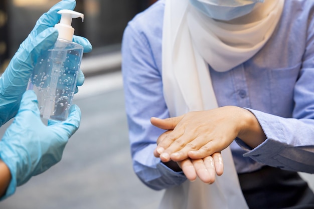 Asiatische Mitarbeiterin oder Ladenbesitzerin trägt Gummihandschuh, während sie Alkoholgel-Händedesinfektionsmittel an muslimische Kunden abgibt Konzept der neuen normalen Geschäftspraxis mit inklusiver Menschenvielfalt