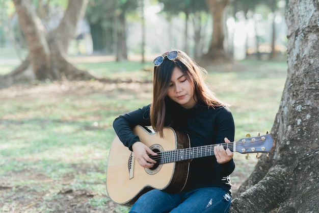 Asiatische Musikerinnen, die ein schwarzes langärmliges Hemd und eine Sonnenbrille auf dem Kopf tragen und morgens unter einem Baum im Park Akustikgitarre singen und spielen