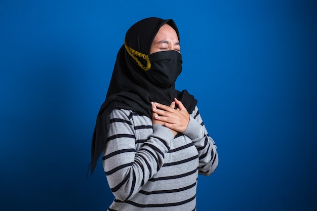 Asiatische muslimische College-Studentin mit Maske, die Schmerzen in der Brust verspürt, Geste, die die linke Brust hält. Blauer Hintergrund