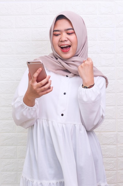 Asiatische muslimische Frau blickt auf ihr Smartphone und erhält gute Nachrichten, die den Sieg zeigen, ja oder gewinnen Geste