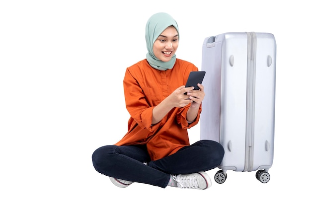 Asiatische muslimische Frau in einem Kopftuch, die mit einem Koffer sitzt, während sie ein Mobiltelefon benutzt