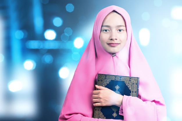 Asiatische muslimische Frau in einem Schleier, der den Koran mit unscharfem hellem Hintergrund steht und hält