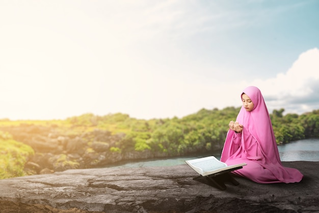 Asiatische muslimische Frau in einem Schleier, der sitzt, während erhobene Hände und im Freien beten