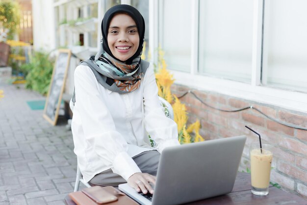 Asiatische muslimische Frau sitzt in einem Garten mit einem Laptop und einem Telefon auf ihrem Schoß