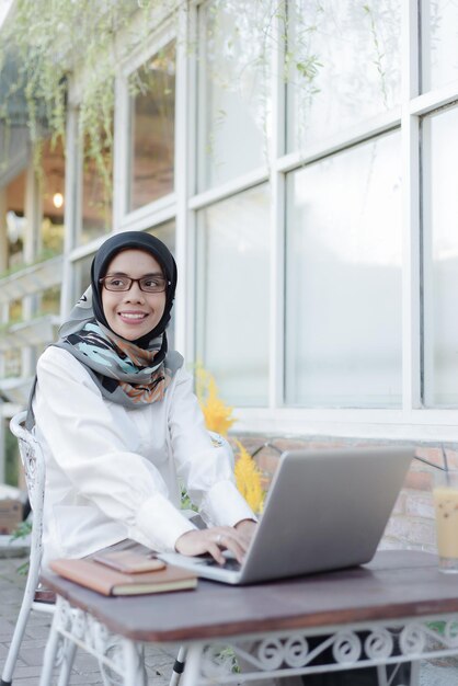 Asiatische muslimische Frau sitzt in einem Garten mit einem Laptop und einem Telefon