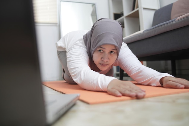 Asiatische muslimische Frau trainiert, beobachtet Online-Video-Unterricht auf einem Laptop und trainiert im Haus