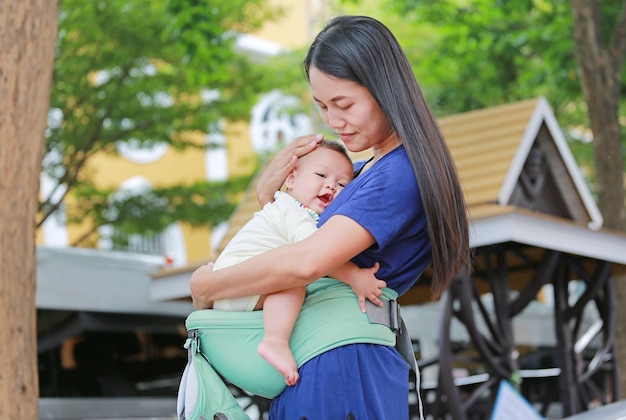 Asiatische Mutter, die ihr Kind auf ergonomischer Babytrage umarmt.