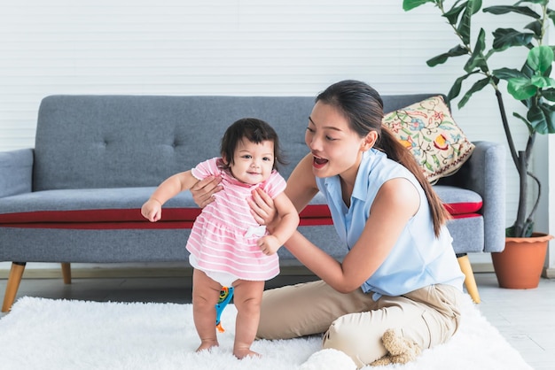 Asiatische Mutter, die lächelt und glücklich ist, während sie ihre 7 Monate alte Tochter unterstützt, die das Stehen lernt