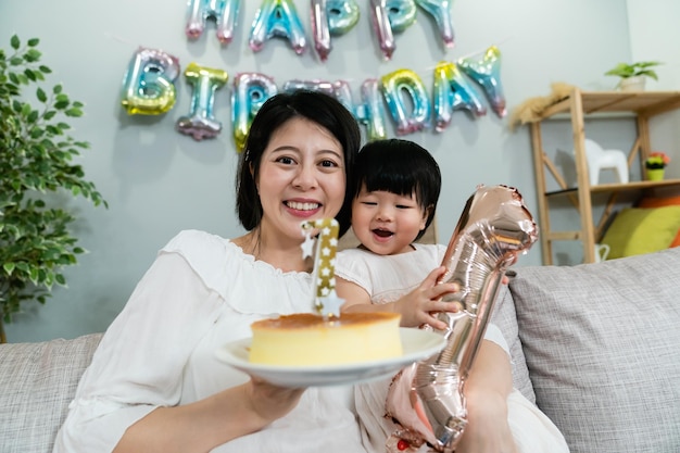 asiatische mutter lächelt in die kamera und hält einen kuchen mit ihrer süßen kleinen tochter in den armen, während sie geburtstag in einem dekorierten wohnzimmer zu hause feiert